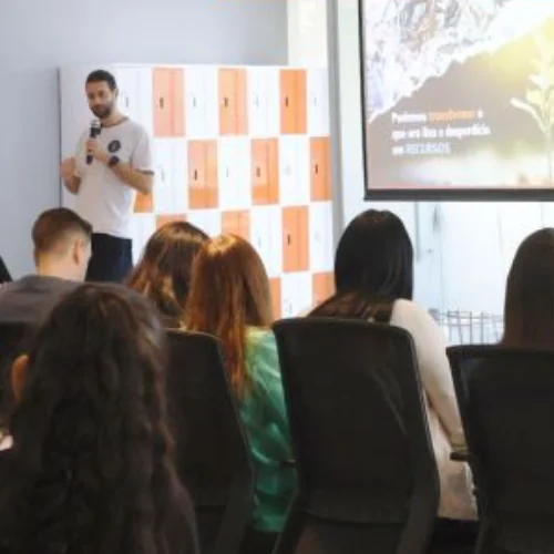 Facilitador apresentando em frente a uma parede de armários laranja e a um telão, para uma plateia sentada